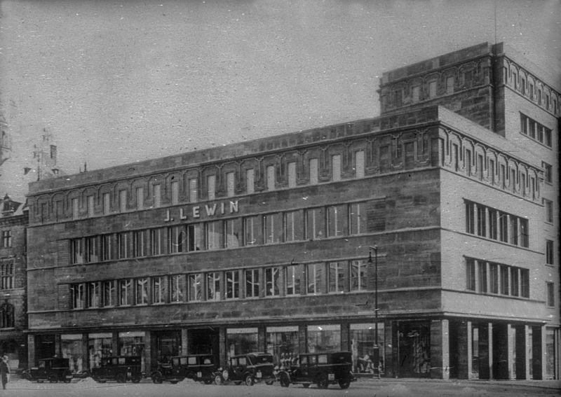 Historisches Schwarz-Weiß-Bild eines vierstöckigen Kaufhauses mit sechsstöckigem Turm, davor mehrere geparkte Autos. An der Fassade steht der Schriftzug J. Lewin.